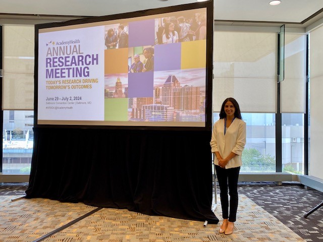 Brynne Presser Funderburg standing by the promotional screen for the 2024 AcademyHealth Annual Research Meeting (Today's Research driving Tomorrow's outcomes.)