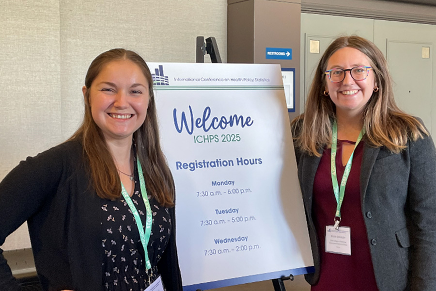 Two women smiling while standing next to a Welcome ICHPS 2025 sign with registration hours
