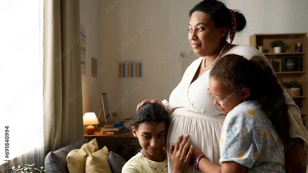 A pregnant women hugging two of her children in their living room while she looks out a nearby window