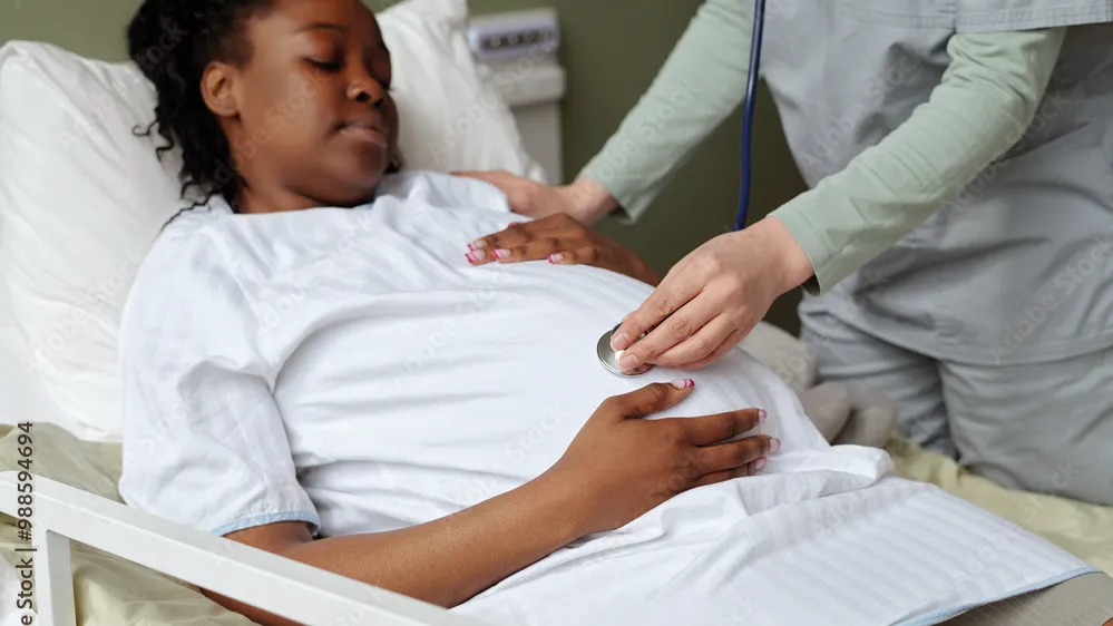 A nurse holding a stethoscope to a pregnant woman's stomach while she lies in a medical bed