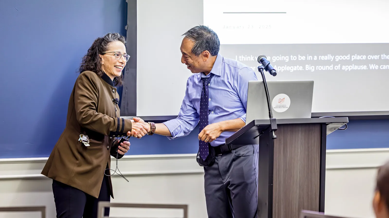 Mary Applegate wearing a brown suit jacket smiling and shaking the hand of Director Liu at the podium.