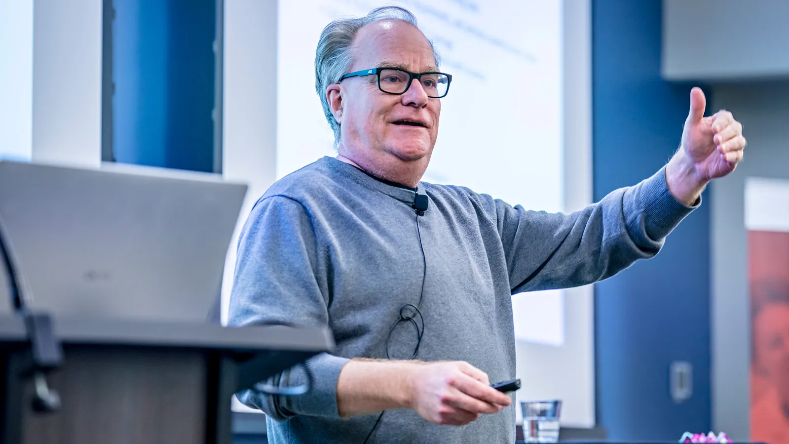 Bill Hayes presenting in front of a podium and screen, raising his hand and holding a clicker.