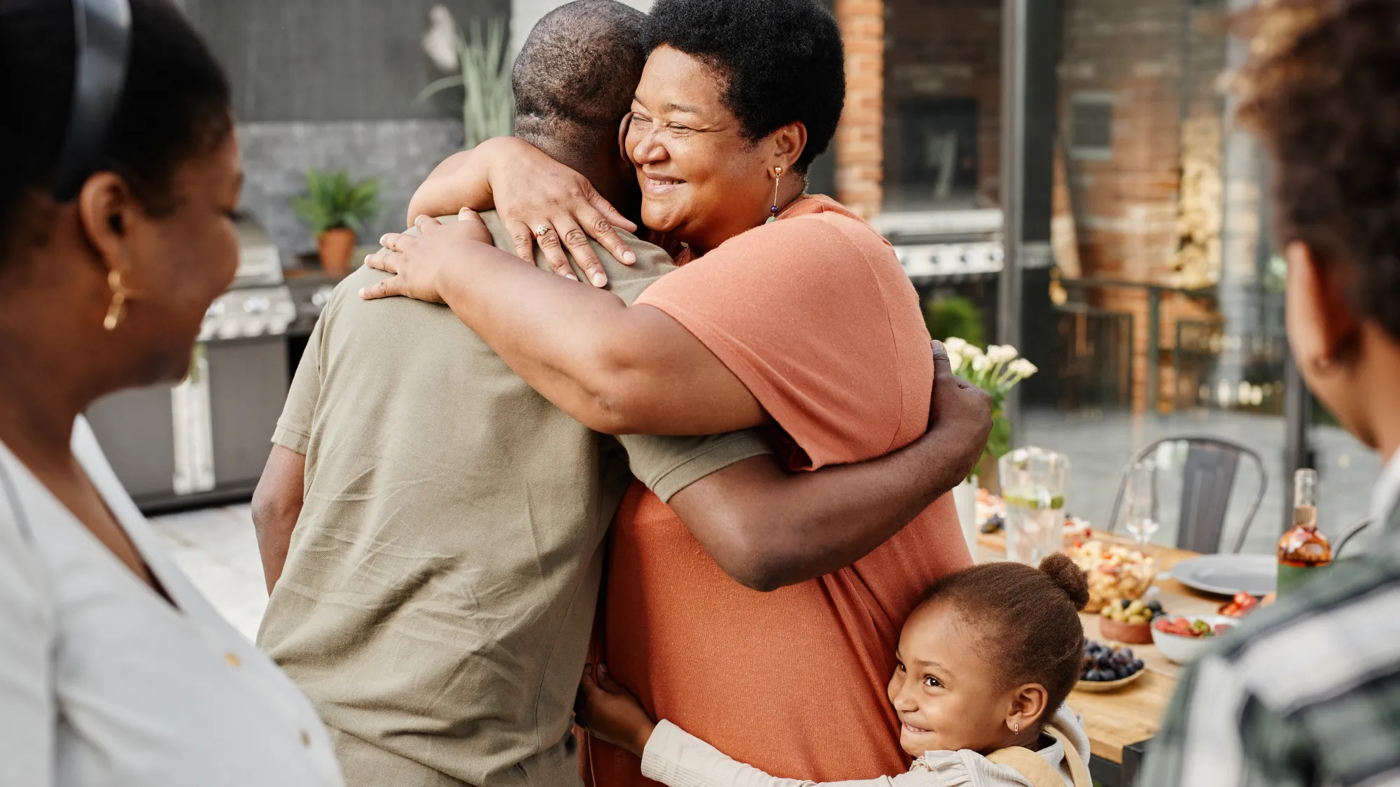 At a barbecue, a smiling woman hugs a man while a child clutches onto her waist