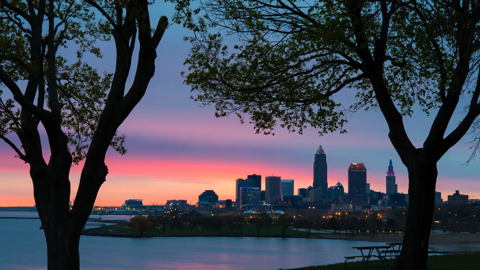 The city of Cleveland taken from a lake Erie beach at dusk.  Has trees, the beach, a picnic table in the foreground with Cleveland in the background
