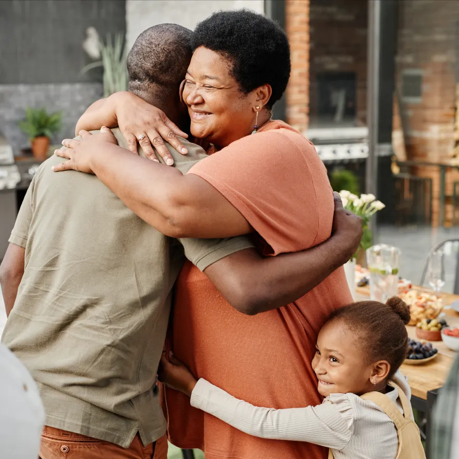 At a barbecue, a smiling woman hugs a man while a child clutches onto her waist